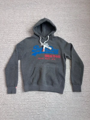Grå Superdry hoodie med tryck - Snygg grå hoodie från Superdry med blått och rött tryck framtill samt vita dragsnören i huvan. Klassisk känguruficka och mjukt material som känns skönt mot huden. Perfekt för dig som gillar streetwear och vill ha en hoodie med coola detaljer.