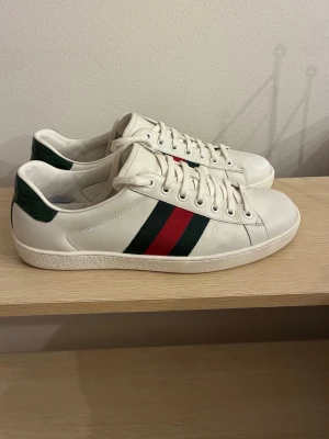 Gucci Ace  - Snygga vita sneakers från Gucci i läder med klassiska gröna och röda sidoremsor samt mörkgrön detalj vid hälen. Skorna har lågt skaft och vita snören. Perfekta för dig som vill ha en clean och lyxig look med ikoniska detaljer.