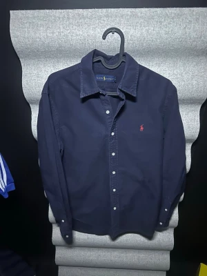 Mörkblå skjorta från Ralph Lauren - Klassisk mörkblå skjorta från Ralph Lauren med knappar framtill och broderad röd logga på bröstet. Skjortan har lång ärm och är tillverkad i bomull, perfekt för en stilren look.