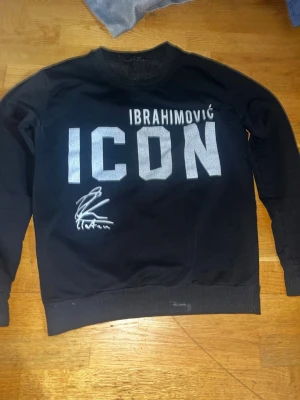 Svart ICON sweatshirt Dsquared2 - Cool svart sweatshirt från Dsquared2 med stor vit text 'ICON' och 'IBRAHIMOVIĆ' på bröstet. Tröjan har även en tryckt autografdetalj. Rund halsringning och ribbade muddar. Perfekt för dig som gillar streetwear och sportig stil.