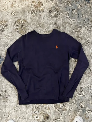 Mörkblå tröja från Ralph Lauren - Mörkblå långärmad tröja från Ralph Lauren med klassisk rund halsringning och den ikoniska röda logotypen broderad på bröstet. Tröjan har ribbade muddar vid ärmslut och nederkant. Perfekt för en stilren och enkel look.