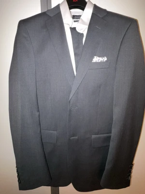 Mörkgrå kostym Dressmann - Snygg mörkgrå kavaj från Dressmann i storlek 44 med slim fit-passform. Kostymbyxor i storlek 30/32. Vit clean skjorta i storlek S. Kläderna är använda 1 gång och är för små för mig. Passar perfekt för någon som är ca 165-170, ca 60-70 kg. 