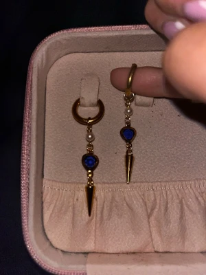 Guldiga örhängen med blå detalj - Säljer ett par trendiga guldiga örhängen med små vita pärlor, blå hjärtformad detalj och spetsig hängande dekoration. Örhängena har en cool och modern design som sticker ut. Perfekta för dig som gillar unika accessoarer med lite edge.