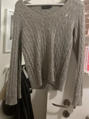 Grå kabelstickad tröja Polo Ralph Lauren - Snygg grå kabelstickad tröja från Polo Ralph Lauren med V halsringning och diskret broderad logga på bröstet. Tröjan har långa ärmar och ribbade muddar. 