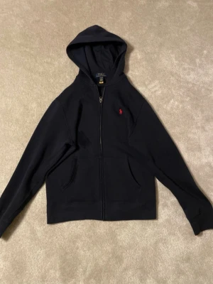 Svart hoodie från Polo Ralph Lauren - Klassisk svart hoodie från Polo Ralph Lauren med dragkedja framtill och huva. Den har två fickor och den ikoniska röda loggan broderad på bröstet. Perfekt för en avslappnad och stilren look. Tillverkad i mjukt bomullsmaterial.