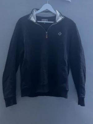 Svart ziptröja (morris) - Snygg svart ziptröja från Morris med halv dragkedja och vit broderad logga på bröstet. Kragen har vita och gröna ränder på insidan och dragkedjan har en läderdetalj. Perfekt till chill vardagsstil.