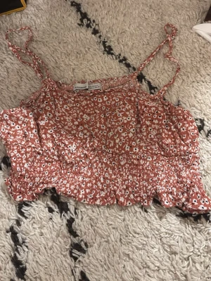 Blommig croppad linnetopp från Hollister - Söt croppad linnetopp från Hollister med smala axelband och blommigt mönster i vitt och rödbrunt. Toppen har smockad midja och volangkant nedtill, perfekt för varma dagar och sommarvibbar.