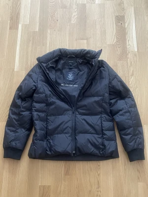 Svart dunjacka från Gant - Svart dunjacka från Gant med quiltad design och hög krage. Jackan har dragkedja framtill och ribbade muddar vid ärmsluten. Perfekt för kyliga dagar och enkel att matcha med olika outfits.