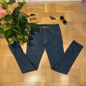 Blå jeans från Nudie Jeans W30 L34 - Säljer ett par klassiska blå jeans från Nudie Jeans i storlek W30 L34. Jeansen har raka ben, fem fickor och snygga kontrastsömmar. Materialet är slitstarkt denim i bomull och passformen är normal. Perfekta för dig som gillar stilrena och tidlösa jeans.