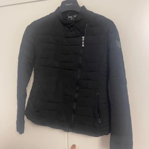 Svart pufferjacka från Hollies - Svart pufferjacka från Hollies med quiltad design och diskret logga på ärmen. Jackan har snedställd dragkedja, två fickor med dragkedja och en hög krage med tryckknappar. Perfekt för kyliga dagar och har en clean, stilren look.