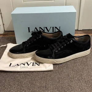  Lanvin sneakers i mocka och lack - Säljer ett par svarta Lanvin sneakers i lyxig kombination av mocka och lackat kalvskinn. Klassisk låg modell med rund tå, svarta snören och vit gummisula. Kommer med originalbox och dustbag. Perfekta för dig som gillar stilrena och exklusiva sneakers.