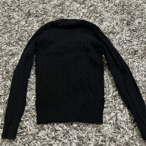 Svart kabelstickad tröja från GANT - Svart kabelstickad tröja från GANT med rund halsringning och broderad logga på bröstet. Tröjan är i bomull och ribbade muddar vid ärmslut och nederkant. Storleken är xs men passar s också