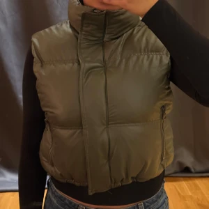 Olivgrön dunväst från FB Sister XS - Säljer en olivgrön dunväst från FB Sister i storlek XS. Västen har hög krage, dragkedja och praktiska fickor med dragkedja på sidorna. Den quiltade designen ger en snygg och puffig look, perfekt att styla över en hoodie eller långärmad tröja.