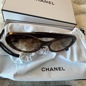 Bruna ovala solglasögon från Chanel - Snygga ovala solglasögon från Chanel i brunmelerad plast med gulddetaljer och kedja längs bågen. Ikonisk design med exklusiv känsla, perfekt accessoar för att lyfta din stil. Levereras med originalfodral och box.