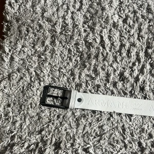 Vit skinnbälte från Armani - Snyggt vitt bälte från Armani i skinn med präglad logotyp längs hela remmen. Bältet har ett stilrent svart metallspänne som ger en modern kontrast till det ljusa lädret. Perfekt accessoar för att lyfta din outfit med en exklusiv touch.