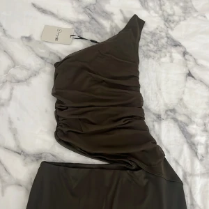 Mörkgrön tajt klänning  - Supersnygg långklänning i mörkgrönt från Design by Si. Klänningen har omlott-design, oneshoulder och tight passform som framhäver figuren. Materialet är mjukt och stretchigt, perfekt för dig som gillar trendiga och unika plagg. Asymmetrisk midja och rynkade detaljer.