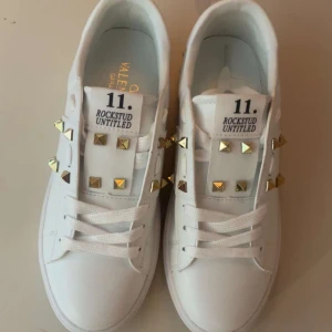 Valentino Rockstud Untitled sneakers vit - Snygga vita Valentino Rockstud Untitled sneakers med ikoniska guldfärgade nitar och klassisk snörning. Skorna är i skinn och har en clean, modern look med coola detaljer som gör dem till ett statement. Perfekta för dig som vill sticka ut med exklusiv streetstyle.