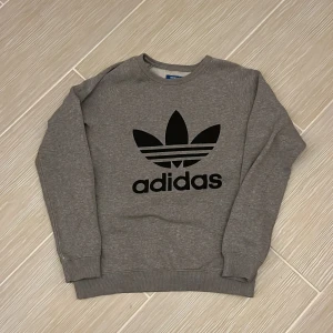Grå sweatshirt från Adidas Classic - Grå sweatshirt från Adidas med stor svart trefoil-logga framtill. Klassisk rund halsringning och ribbade muddar vid ärmslut och nederkant. Tillverkad i mjuk bomullsblandning, perfekt för en chill och sportig look.