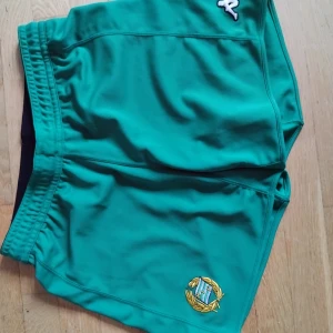 Gröna sportshorts XL med brodyr - Säljer ett par gröna sportshorts i storlek XL med broderad logga och elastisk midja med snörning. Shortsen är tillverkade i 100% polyester och har en klassisk sportig look, perfekta för träning eller fotboll.