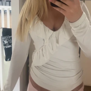 Vit långärmad blus med volang - Supersöt vit blus med långa ärmar och djup v-ringning. Toppen har en snygg volangdetalj som går längs ringningen och snett över bröstet. Figurnära passform och mjukt material som känns skönt mot huden. Perfekt för dig som gillar romantiska och trendiga plagg.