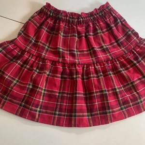 Rutig röd kjol från H&M - Supersöt röd kortkjol från H&M med klassiskt rutigt mönster i svart, vitt och gult. Kjolen har resår i midjan och volangdetalj som ger extra volym. Perfekt för dig som gillar färg och mönster!