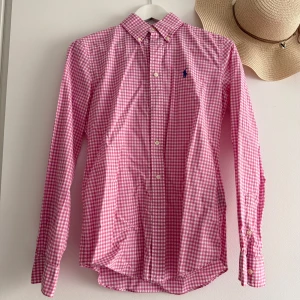 Rosa rutig skjorta från Ralph Lauren - Säljer en snygg rosa och vit rutig skjorta från Ralph Lauren med klassisk krage och lång ärm. Skjortan har custom fit-passform, knappar framtill och ikonisk blå logga broderad på bröstet. Perfekt för dig som gillar preppy stil.