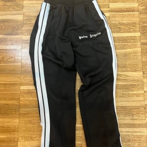 Palm Angels svarta trackpants med ränder - Svarta trackpants från Palm Angels med vita sidoränder och logga broderad på vänster sida. Byxorna har elastisk midja och är tillverkade i ett glansigt syntetmaterial. Perfekta för en sportig och trendig streetwear-look.