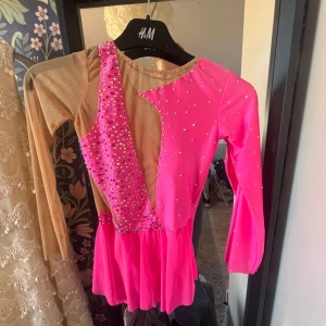 Glittrig rosa konståkningsklänning💕🩷🤩 - Säljer en supercool kort konståkningsdräkt i starkt rosa med glittriga stenar och nude meshdetaljer. Klänningen har långärmade meshärmar och asymmetrisk design framtill. Perfekt för dig som vill sticka ut på isen!🩷🫶🏻Bara använd två gånger😇🥰