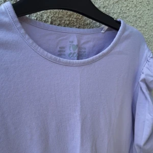 Ljuslila och ljusrosa långärmade toppar - Två långärmade toppar från I Love Eco, en i ljuslila och en i ljusrosa. Båda har rynkade detaljer längs ärmarna och rund halsringning. Den ljusrosa toppen är ribbad medan den lila är slät. Perfekta för vår och höst!