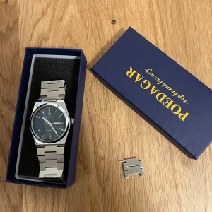 Silverfärgad klocka från Poedagar - Stilren klocka från Poedagar med silverfärgat armband i metall och svart urtavla. Klockan har dag- och datumvisning samt klassisk länkdesign. Perfekt klocka för dig som gillar en modern och tidlös look.