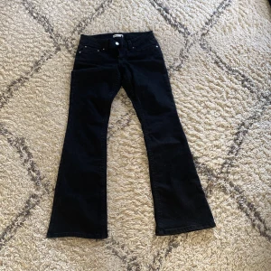 Svarta bootcut jeans med fickdetaljer - Svarta jeans med bootcut passform och snygga fickor med lock och knappar bak. Klassisk femficksmodell framtill och bälteshällor. Jeansen har en stilren look och är tillverkade i ett mjukt denimtyg som sitter skönt.