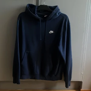 Mörkblå Nike hoodie med logga - Snygg mörkblå hoodie från Nike med klassisk vit logga på bröstet. Tröjan har huva med snörning och magficka. Perfekt för dig som gillar en avslappnad och sportig stil. Hoodien är i storlek S men kan även passa M.