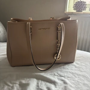 Beige handväska från Michael Kors - Stilren beige handväska från Michael Kors i skinn med guldiga metalldetaljer och MK-logga. Väskan har två långa handtag, flera fack inuti och dragkedja i mitten. Perfekt för dig som vill ha en klassisk och rymlig väska med exklusiv känsla. Några små fläckar inombords som ej syns 