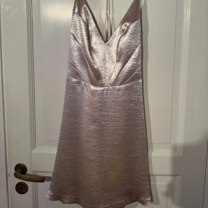 Champagnefärgad kort klänning med v-ringning - Säljer en glansig champagnefärgad kort klänning med v-ringad front och tunna axelband. Klänningen har en lätt A-linjeformad siluett och är tillverkad i ett skimrande satinliknande material som ger en lyxig känsla. Perfekt för dig som vill sticka ut med en elegant look.