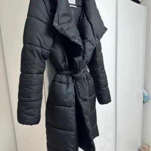 Svart lång pufferjacka med huva - Säljer en svart, lång pufferjacka med huva och bälte i midjan. Jackan har quiltad design, långa ärmar och en snygg oversized krage. Perfekt för kalla dagar och har en riktigt chill vibe. Bältet gör att du kan justera passformen enkelt.