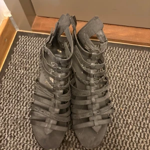 Svarta gladiatorsandaler från Duffy - Svarta gladiatorinspirerade sandaler från Duffy i storlek 40. Skorna har flera remmar, flätade detaljer och dragkedja bak för enkel påtagning. Platt sula och öppen tå ger en cool och luftig känsla. Perfekta för varma dagar och snygga till både shorts och klänning.