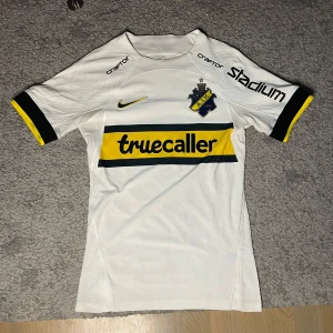 AIK fotbollströja  - AIK:s officiella vita matchtröja från Nike med Salétros och nummer 7 på ryggen. Tröjan har svarta och gula detaljer, AIK-emblem på bröstet och sponsorer som Truecaller och Stadium. Tillverkad i lätt och ventilerande material, perfekt för fotbollsplanen. Storlek XS