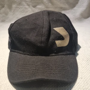 Svart keps från Kolla Kolla - Svart keps med justerbar snapback och ett stort guldigt tryck framtill. Klassisk form med böjd skärm och ventilerande hål upptill. Tillverkad i bomullstyg, perfekt för en streetwear-look.