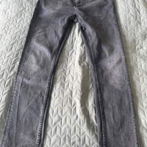 Grå skinny jeans från Nudie Jeans - Säljer ett par grå skinny jeans från Nudie Jeans med klassisk broderad bakficka och svarta detaljer. Jeansen har en smal passform och är tillverkade i slitstarkt denim. Perfekta för dig som gillar en clean och stilren look.