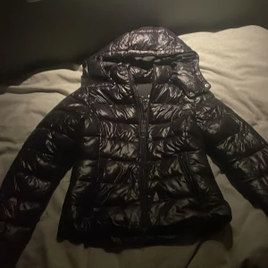 Svart pufferjacka från 157, XS - Svart glansig pufferjacka från 157 i storlek XS. Jackan har huva, dragkedja framtill.
