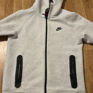 Ljusgrå Nike hoodie med dragkedja - Säljer en ljusgrå hoodie från Nike med hel dragkedja framtill och svarta detaljer. Tröjan har huva, två fickor med svarta dragkedjor och Nike-logga på bröstet. Perfekt för dig som gillar sportig och clean stil.