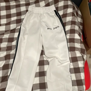 Vita Palm Angels trackpants med svarta ränder - Säljer ett par vita trackpants från Palm Angels med svarta sidoränder och broderad logga på framsidan. Byxorna har elastisk midja med snörning och raka ben. Perfekta för en streetwear-look och riktigt sköna att bära.