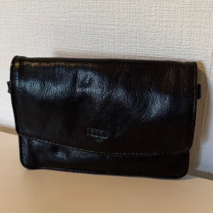 Axelremsväska - Stilren svart väska från Ceannis i äkta skinn. Plånboken/väskan har flera fack, dragkedja på baksidan och magnetknäppning. Perfekt för att hålla ordning på kort, kontanter och småprylar. Klassisk design som funkar till allt.