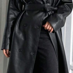 Svart skinn trenchcoat med bälte - Snygg svart trenchcoat i skinnimitation med breda slag, knappar framtill och matchande bälte i midjan. Jackan har lång ärm och en clean, minimalistisk look som ger en cool vibe. Perfekt för dig som vill sticka ut med en edgy stil. Endast använd en gång