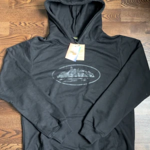 Corteiz Hoodie - Svart hoodie med ett coolt tryck av en byggnad i grått på bröstet. Klassisk huva, känguruficka och ribbade muddar. Perfekt för dig som gillar streetwear och vill ha en stilren look.