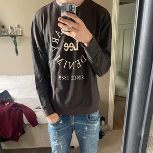 Svart sweatshirt från Lee med tryck - Svart sweatshirt från Lee med stort vitt tryck på bröstet där det står 'Legendary Denim Since 1889'. Tröjan har rund hals, långa ärmar och en relaxed passform. Mjuk insida och klassisk stil som funkar till jeans. Bra skick, lite urtvättad men inga övriga fel med tröjan. Hör av dig om du har några funderingar