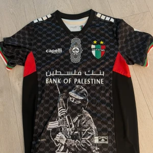 Palestino T-Shirt  - Unik Palestino fotbollströja från Capelli Sport i svart med grått mönster, röda detaljer och klubbmärke på bröstet. Tryck med Bank of Palestine och illustration framtill. Korta ärmar med ränder i grönt, vitt och rött. Tillverkad i lätt polyester för maximal komfort.