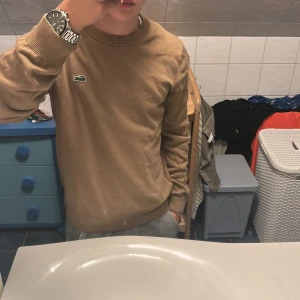 Beige tröja från Lacoste i bomull - Säljer en beige tröja från Lacoste med klassisk rund hals och den ikoniska krokodilloggan på bröstet. Tröjan är i bomull och har ribbade muddar vid ärmslut och nederkant. Regular fit och långärmad, perfekt för en clean och stilren look. Mycket fint skick utan defekter!