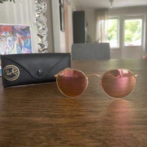 Ray-Ban Round Metal solglasögon guld - Säljer ett par Ray-Ban Round Metal solglasögon med tunna guldfärgade metallbågar och runda linser i rosa ton. Modellen är RB3447 och kommer med original Ray-Ban-fodral. Säljer då de ej passar min ansiktsform. Superfina!