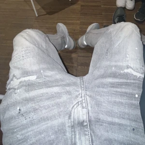 Grå slitna jeans - Säljer ett par grå jeans från Supply & Demand med slitningar, hål och vita färgstänk för en riktigt edgy look. Jeansen har fem fickor, normal passform och raka ben. Perfekta för dig som gillar streetstyle och vill sticka ut.